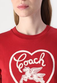 Camiseta de algodón roja con un diseño de corazón blanco que incluye "Coach" y un gráfico de un caballo alado, con escote redondo y mangas cortas.