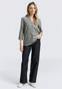 Graue karierte Bluse mit V-Ausschnitt, Dreiviertelärmeln und asymmetrischem Saum, kombiniert mit dunkler Jeanshose und weißen Sneakers.