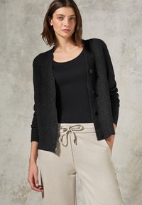 Zwarte gebreide cardigan met een gestructureerde afwerking en knoopsluiting, gedragen over een zwart topje en lichtbeige wijde broek.
