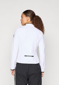 Valge pika varrukaga spordijakk kõrge kaelusega, mustade aksentidega mansettide ja ääres ning logoga seljal. Sile ja veniv materjal.