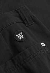 Zwarte broek met visgraatstructuur, voorzien van een ronde patch met "A" logo en een drukknoopdetail op de zak.