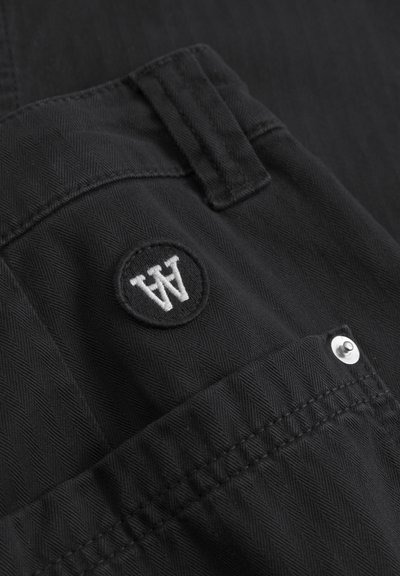 Musta housut, joissa on herringbone-kuvio, ja niissä on pyöreä logo-patch, jossa on "A"-logo, sekä nappikiinnitys taskussa.