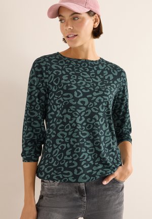 Maglia verde a maniche lunghe con stampa leopardata nera, scollo rotondo e tessuto morbido. Modella abbinata a jeans grigi chiari e a un cappellino di corduroy rosa.