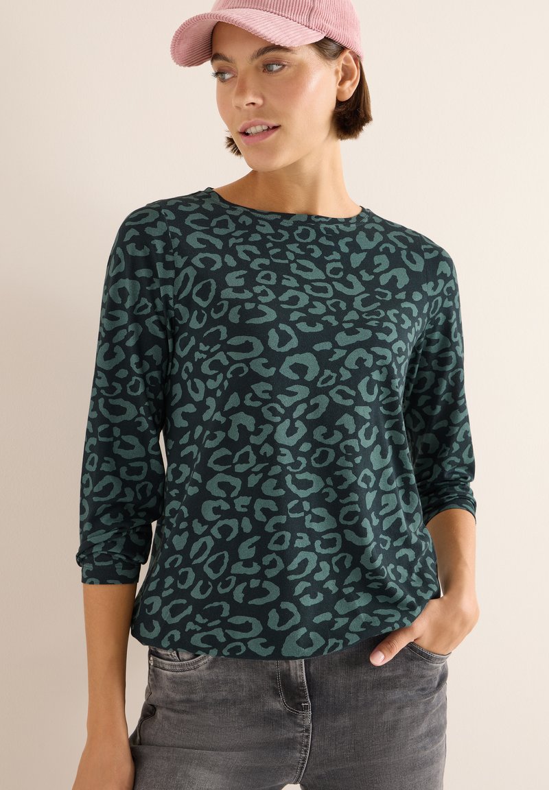 Grünes Longsleeve-Oberteil mit schwarz-leopardenmuster, runder Ausschnitt und weichem Stoff. Modell kombiniert mit hellgrauen Jeans und einer pinken Cordmütze.