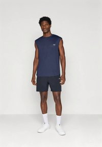 Mężczyzna stoi w granatowej bezrękawnik, dopasowanych szortach, białych skarpetkach sportowych i białych butach sportowych na tle jednolitego jasnego tła.