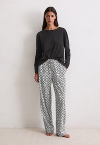 Mujer de pie descalza, vestida con una blusa negra de manga larga y pantalones sueltos blancos con estampado, con la mano en el bolsillo, frente a una pared lisa.
