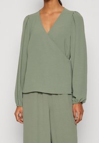 Top avvolgente verde chiaro in tessuto testurizzato, con maniche lunghe arricciate e scollatura a V profonda, abbinato a pantaloni ampi coordinati.
