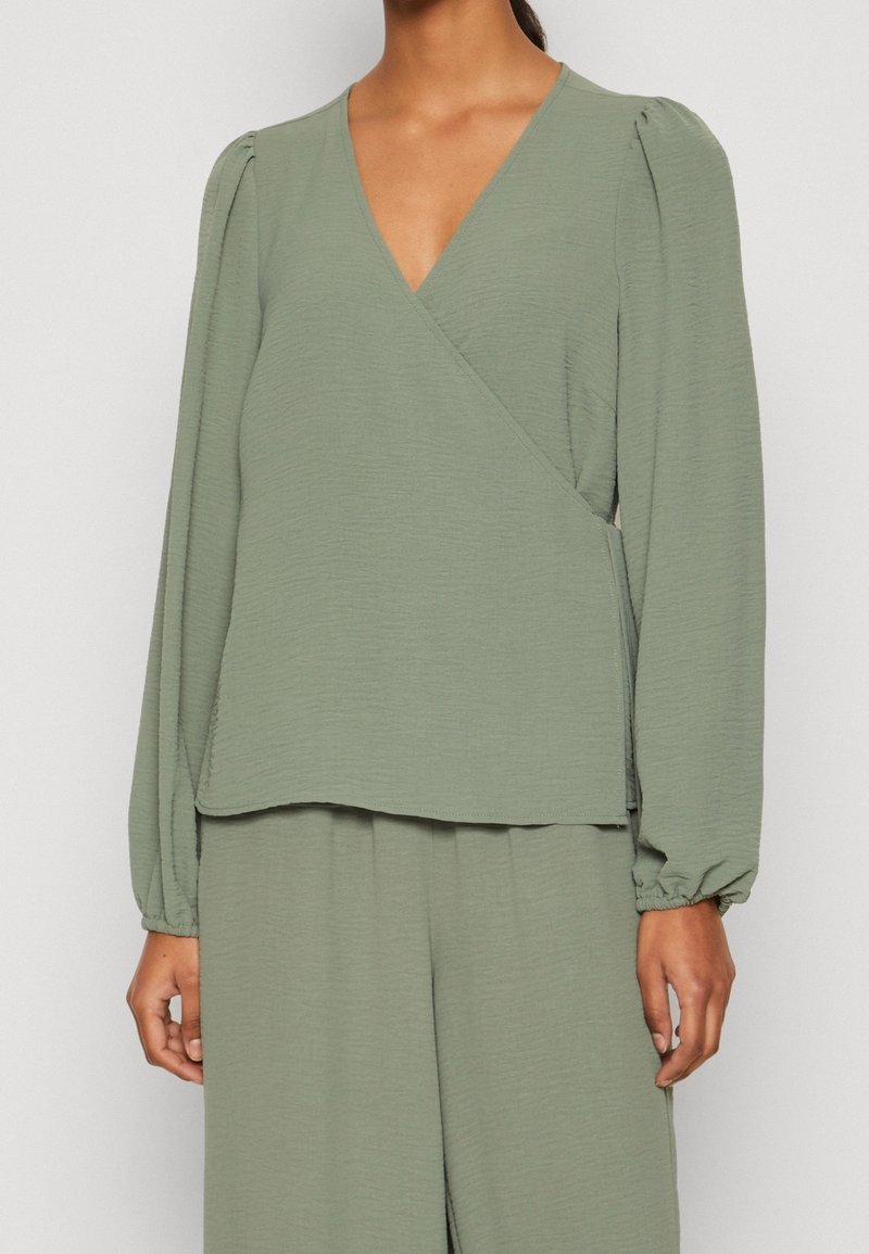 Top avvolgente verde chiaro in tessuto testurizzato, con maniche lunghe arricciate e scollatura a V profonda, abbinato a pantaloni ampi coordinati.