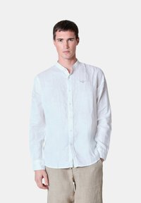 Camicia bianca di lino a maniche lunghe, con bottoni frontali, colletto alla mandarino, dettagli ricamati discreti sul petto, abbinata a pantaloni beige.