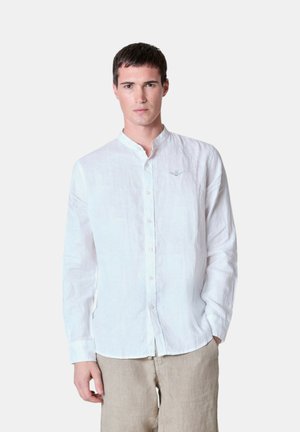 Camicia bianca di lino a maniche lunghe, con bottoni frontali, colletto alla mandarino, dettagli ricamati discreti sul petto, abbinata a pantaloni beige.