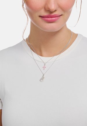 Silberne Halsketten in Lagen, mit einem rosa Kreuzanhänger und einem herzförmigen Design, getragen über einem schlichten weißen T-Shirt.