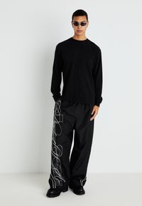 Camisa negra de manga larga con un corte relajado, combinada con pantalones negros de pierna ancha que presentan diseños de líneas abstractas blancas y zapatos planos negros.