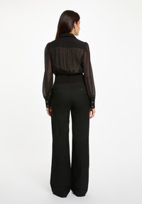 Blusa nera con retro trasparente e plissettato e vita aderente; abbinata a pantaloni neri a gamba larga a vita alta con dettagli a bottoni.