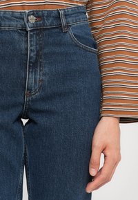 Høyt liv blå denim jeans med glidelås og metallknapp, med minimal søm og en glatt tekstur. Stripete brun topp synlig.