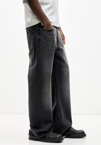 Pantalones vaqueros anchos negros fabricados en denim, con un acabado desgastado, bolsillos laterales y detalles de costura estándar. Combinados con zapatos negros.