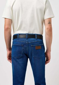 Donkerblauwe spijkerbroek met een rechte snit, voorzien van een zwarte leren riem, twee achterzakken en een bruine leren merkpatch.