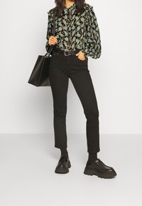 Blusa nera con motivo a fantasia, decorata con un design floreale blu e arancione, abbinata a pantaloni neri slim-fit e scarpe nere stringate chunky, mentre tiene una borsa tote nera.