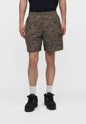 Homme portant un short vert olive à motifs avec une taille élastique, chaussures noires, chaussettes blanches, debout devant un fond blanc uni.
