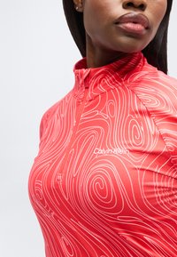 Γυναίκα που φοράει κόκκινο κορυφαίο Calvin Klein με μακριά μανίκια, διακοσμημένο με λευκά κυματιστά μοτίβα και έναν μικρό φερμουάρ μπροστά, πάνω σε απλό φόντο.