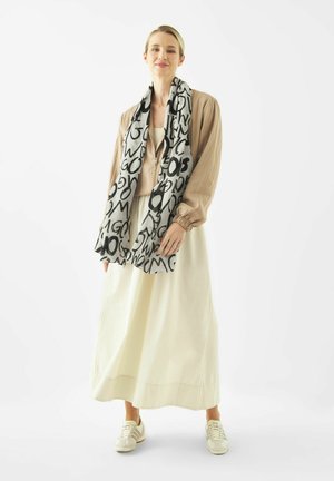 Frau trägt eine beige Jacke, einen weißen langen Rock, weiße Turnschuhe und einen schwarz-weißen Schal mit abstraktem Schriftmuster.