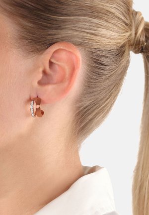 Orecchino ad anello attorcigliato in oro rosa con piccoli cristalli trasparenti incastonati, indossato all'orecchio sinistro di una persona con i capelli biondi raccolti.