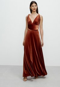 Maxi dress in rust velvet, featuring a deep V-neck, wide straps, and a fitted waist that flows into a flared skirt. Smooth texture, elegant design.