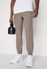Lysbrune Nike-joggers med elastisk afslutning, et lille sølvlogo og en glat struktur. Modellen holder en sort kettlebell og har hvide sneakers på.