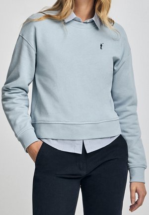 Femme portant un sweat-shirt bleu clair par-dessus une chemise à col avec un petit logo joueur de polo noir sur la poitrine et un pantalon foncé, main dans la poche.