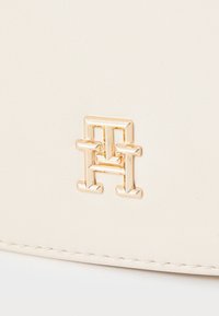 Una borsa a tracolla in pelle color crema con un emblema e logo in tonalità oro e dettagli di cucitura fine. Il materiale ha una superficie liscia.