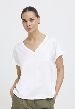 Femme avec les cheveux attachés en chignon, portant des boucles d'oreilles dorées, un chemisier blanc ample à col en V, et un pantalon vert olive, les mains dans les poches.