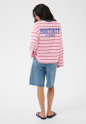 Femme aux longs cheveux foncés portant une chemise rose à rayures avec le texte "Positivity is Power", un short en jean et des tongs bleu marine, de dos.