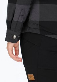 Camicia di flanella a scacchi neri e grigi con polsini bottonati, abbinata a pantaloni neri con una toppa con logo beige sulla coscia.