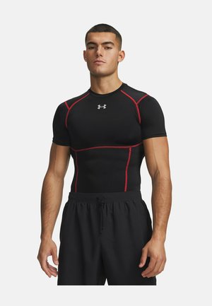 Jonge man die een zwart compressieshirt met rode naden en zwarte shorts draagt, staand tegen een effen witte achtergrond.