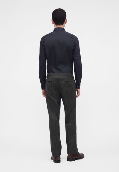 Homme debout de dos, portant une chemise sombre à manches longues, un pantalon gris à fines rayures et des chaussures habillées marron sur fond blanc.