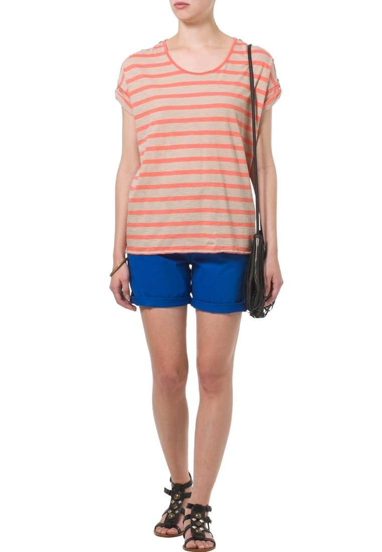 Pruhovaná, krátká béžová košile se světle korálovými pruhy, spárovaná s modrými šortkami. Černé gladiátorské sandály a taška přes rameno.