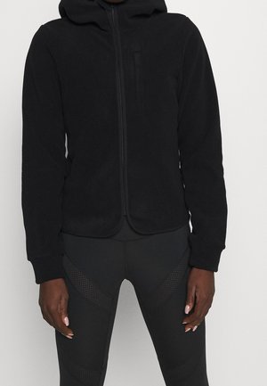Personne portant un sweat à capuche zippé noir et un legging noir avec un détail en mesh, debout devant un fond clair uni.