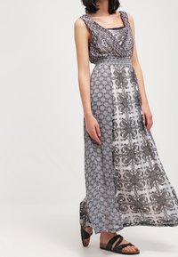 Sukienka maxi z dopasowaną, wzorzystą górą i zwiewnym dołem. Posiada czarno-białe, skomplikowane wzory, krótkie rękawy i talię ściągniętą w pasie.