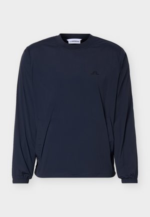 Μπλε σκούρο μπλουζάκι με μακριά μανίκια και στρογγυλή λαιμόκοψη, με μικρό λογότυπο στο στήθος και ελαστικές μανσέτες, παρουσιάζεται σε απλό φωτεινό φόντο.