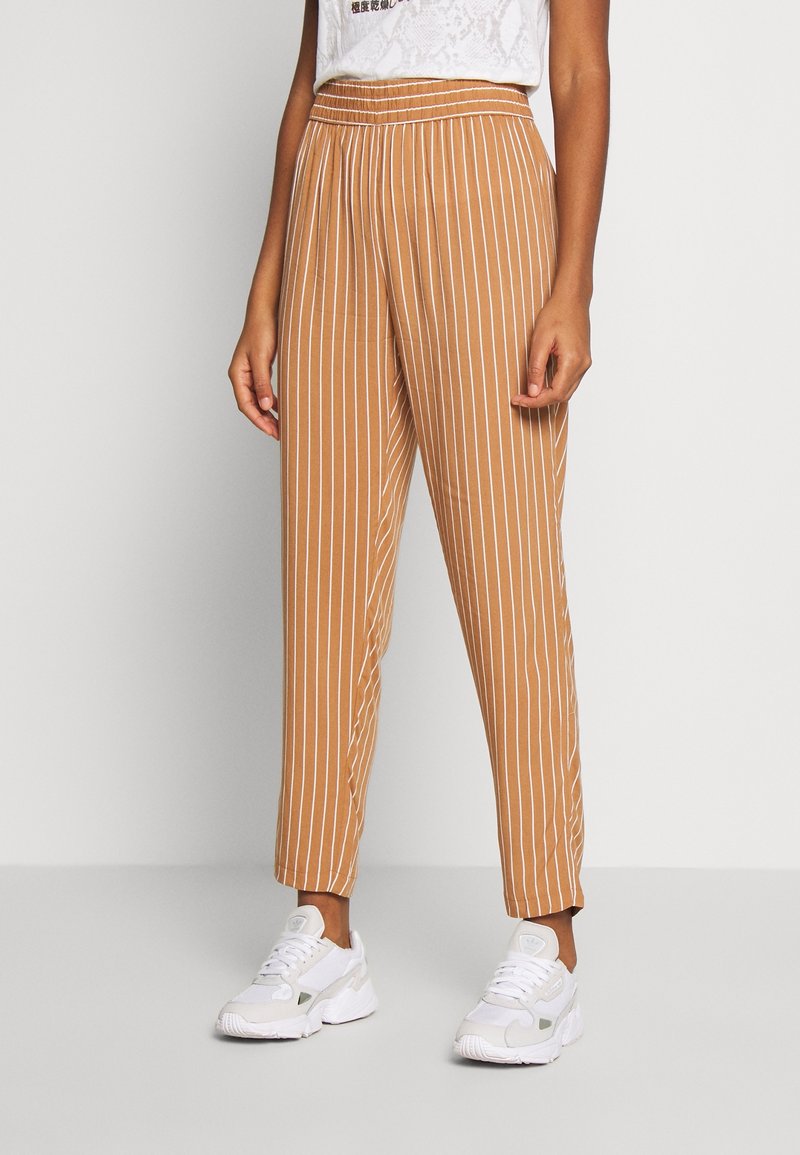 Hnědé kalhoty s vertikálním pruhem a elastickým pasem, s manžetami na kotnících. V kombinaci s bílými teniskami s texturovaným povrchem.