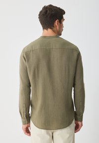 Olive green long-sleeve shirt made of a textured fabric, featuring a rounded hem and buttoned cuffs, shown from the back.