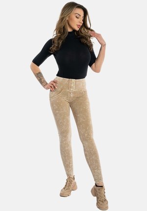 Pantaloni in corda beige a vita alta con chiusura a cinque bottoni, abbinati a una maglietta nera attillata a maniche corte, indossati con scarpe da ginnastica beige con lacci.