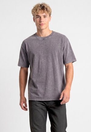 Giovane uomo con capelli biondi ricci che indossa una maglietta grigia slavata oversize e pantaloni neri, in piedi davanti a uno sfondo chiaro e uniforme.