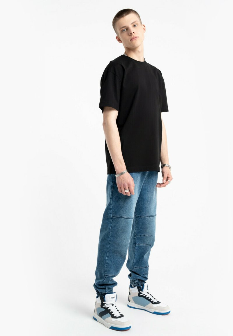 Czarna koszulka z krótkim rękawem zestawiona z niebieskimi dżinsowymi joggerami z detalami w postaci przeszyć oraz białymi trampkami z niebieskimi i szarymi akcentami.