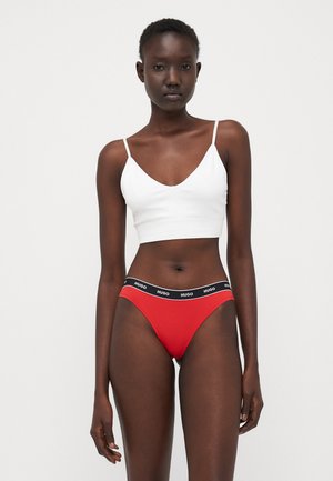Witte cropped tanktop met dunne banden, gecombineerd met rode bikini briefs met een zwarte band die "HUGO" vermeldt. Glad stof, aansluitend ontwerp.