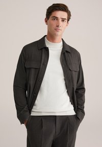 Hombre joven con las manos en los bolsillos, viste una chaqueta oscura abotonada sobre un suéter blanco y pantalones oscuros a juego, contra un fondo liso.