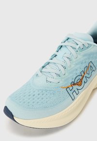 Lichtblauwe hardloopschoen met een gestructureerde mesh bovenkant, een gevoerde tong en veters. Heeft opvallende 'HOKA' branding in oranje, met een contrasterende marineblauwe zool.