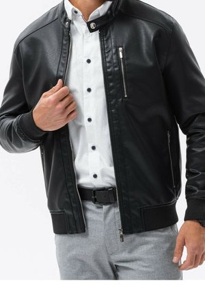 Homme portant une veste en cuir noire sur une chemise blanche boutonnée, pantalon gris et ceinture noire, main ajustant la fermeture éclair de la veste.