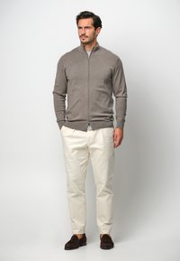 Cardigan zip-up marrone in lana, abbinato a pantaloni color crema e mocassini marrone scuro; texture liscia, design aderente.