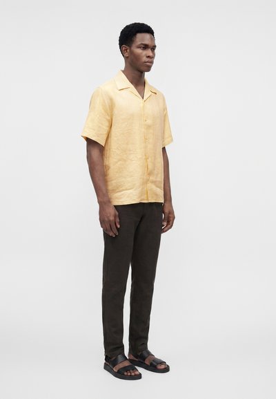 Homme debout portant une chemise beige à manches courtes boutonnée, un pantalon foncé et des sandales noires à bout ouvert sur un fond uni.