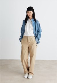 Veste en denim bleu, t-shirt blanc, pantalon beige et baskets blanches. Coupe ample avec des détails de boutons. Textures lisses, couleurs neutres.